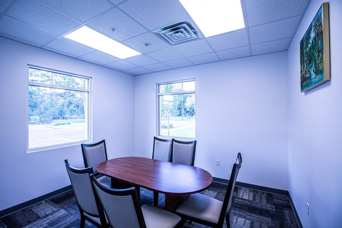 A small conference room with an oval table and six chairs, illuminated by overhead lighting and natural light from two windows.