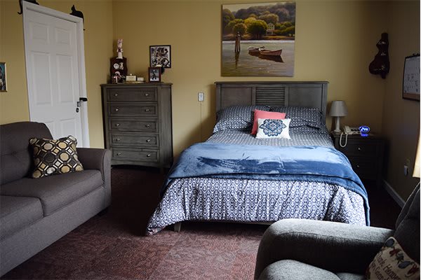 A cozy bedroom with a bed, dresser, and armchair, featuring a landscape painting on the wall.