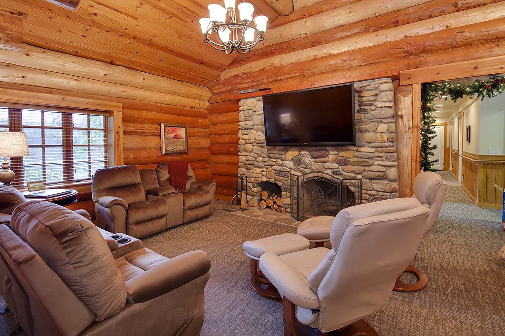 A cozy log cabin living room featuring a stone fireplace, comfortable seating, and a large television.