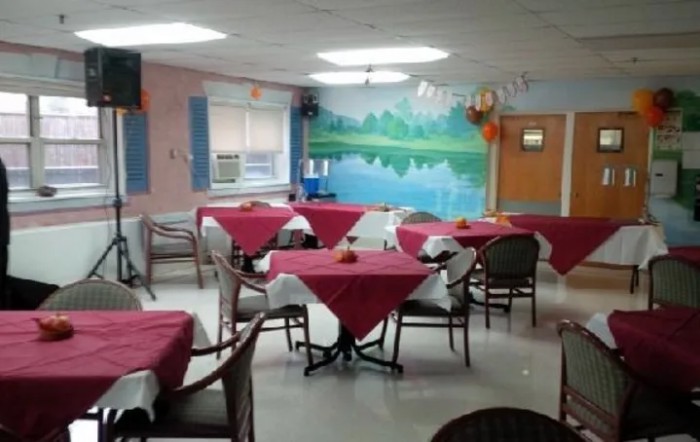 A cozy dining area with tables covered in red tablecloths, surrounded by wicker chairs and featuring a wall mural of a lakeside scene.