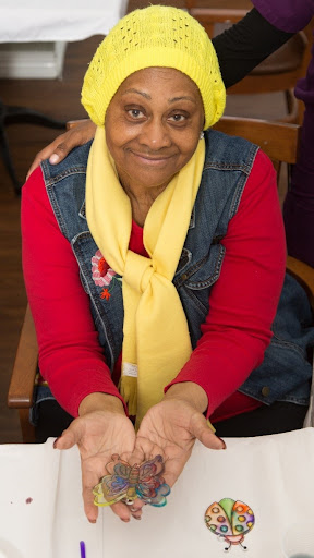 An elderly woman smiles warmly while showcasing her hands, dressed in a bright red shirt with a yellow scarf and denim vest.