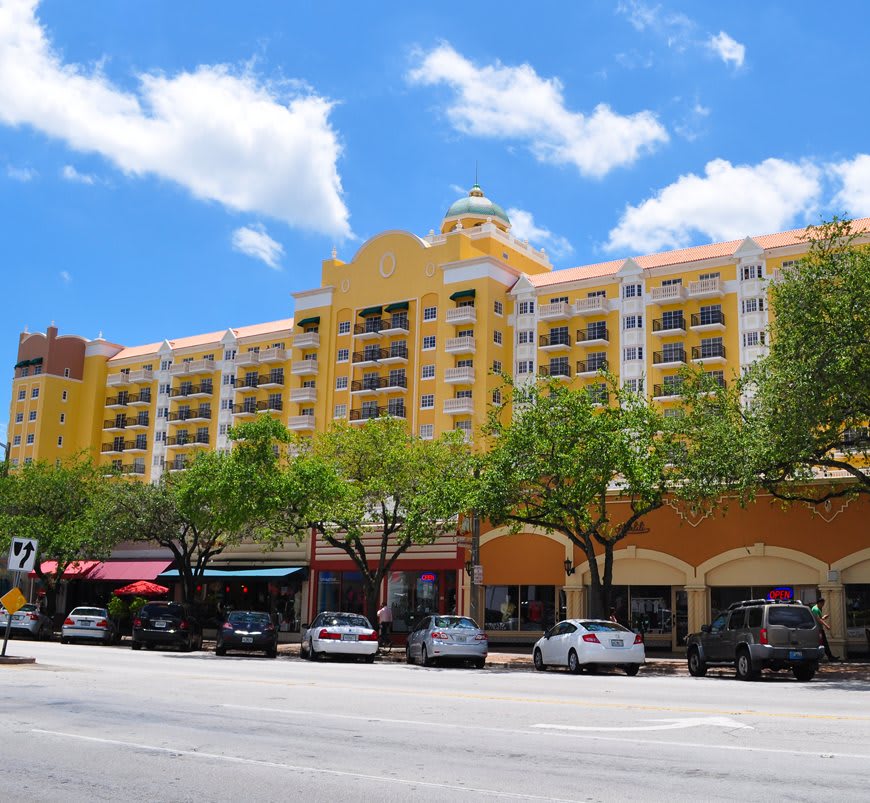 A view of a colorful residential building with shops below, lined with parked cars and trees on a sunny day.