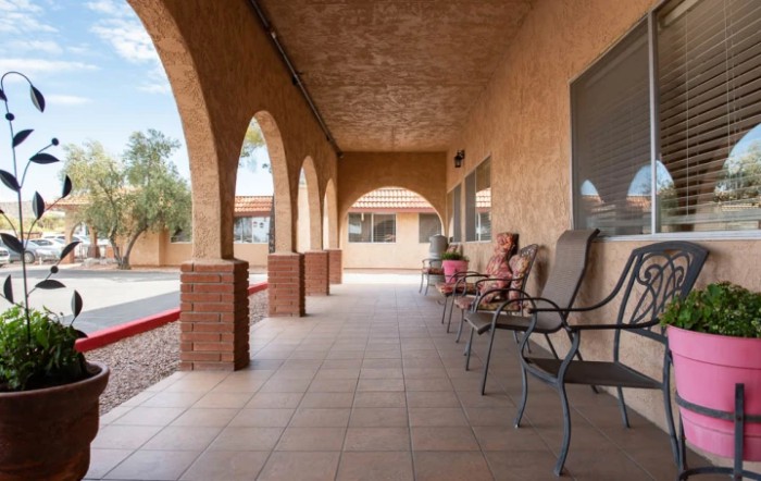 A covered porch with arches, featuring several chairs along the side of a brick building.