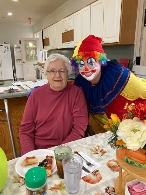 An elderly woman sitting at a dining table beside a smiling clown in a colorful costume.
