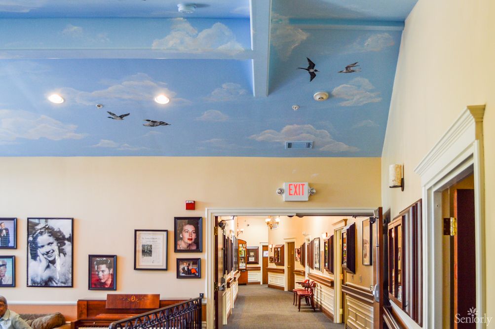 A hallway with a blue sky ceiling mural featuring clouds and birds, adorned with framed paintings along the walls.