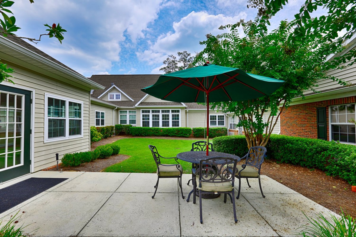 A cozy outdoor patio area with a table, chairs, and an umbrella surrounded by greenery.