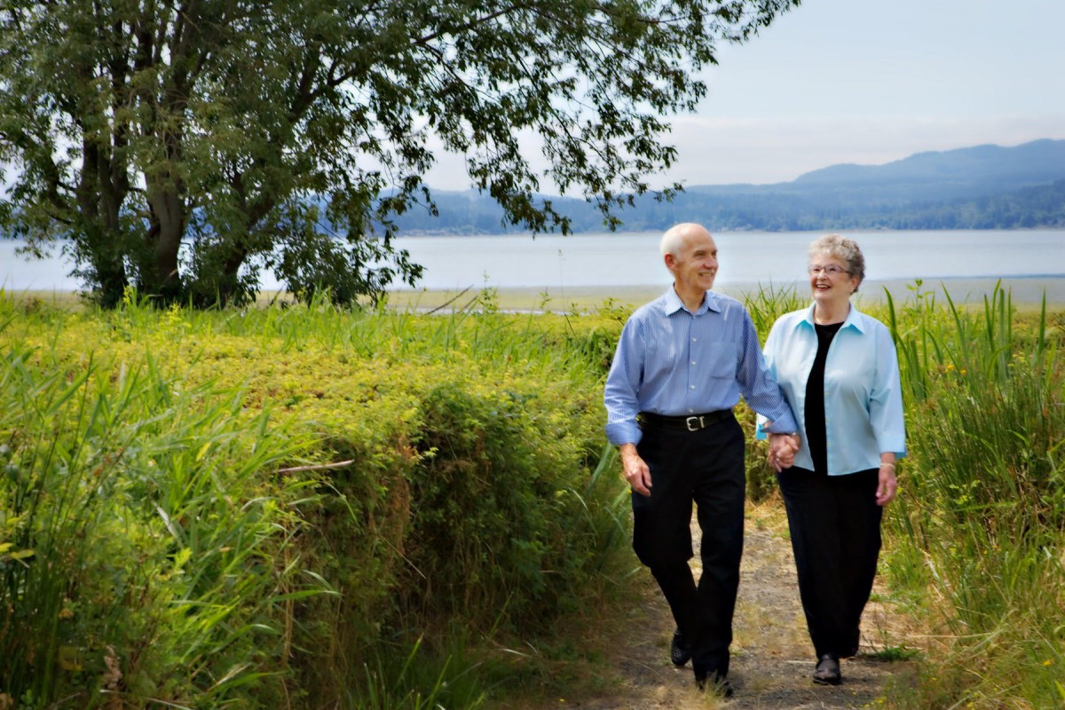 An elderly couple walking hand in hand along a scenic lakeside pathway.