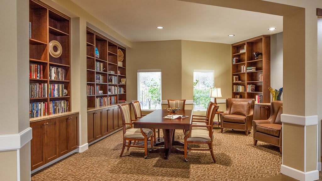 A cozy library space with bookshelves, a central table surrounded by chairs, and warm lighting.