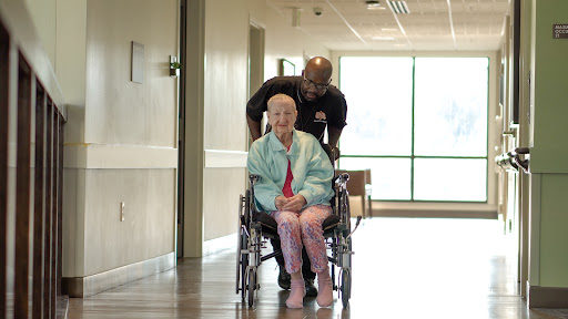 A caregiver gently pushes an elderly woman in a wheelchair down a bright hospital hallway.