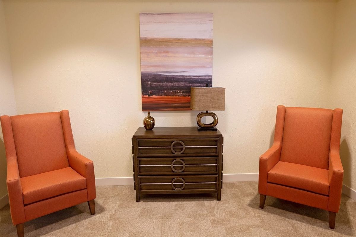 A cozy living room setup featuring two orange chairs flanking a wooden chest with a lamp and decorative items, beneath an abstract painting.