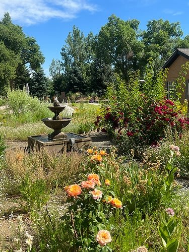 A vibrant garden with a three-tiered fountain surrounded by blooming flowers and lush greenery under a clear blue sky.