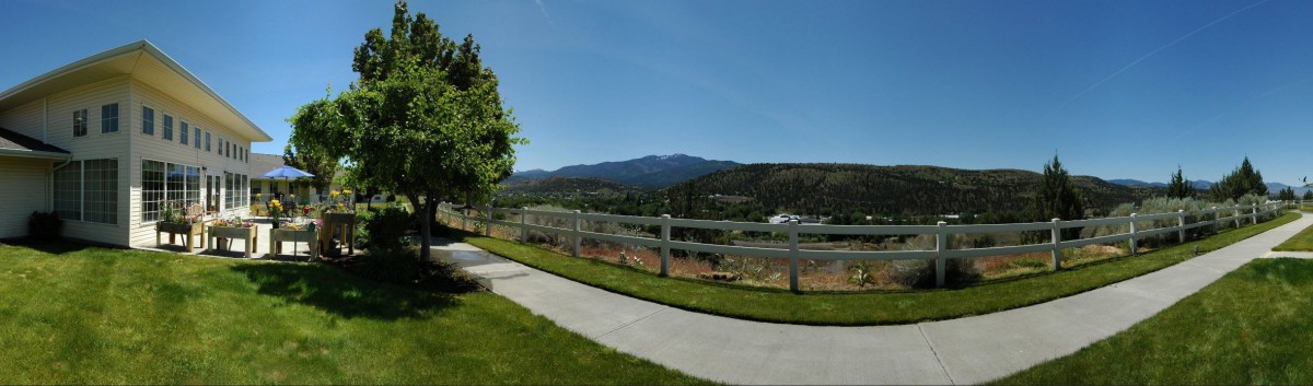 A scenic view featuring mountains in the background, a white fence along a pathway, and lush green grass under a clear blue sky.