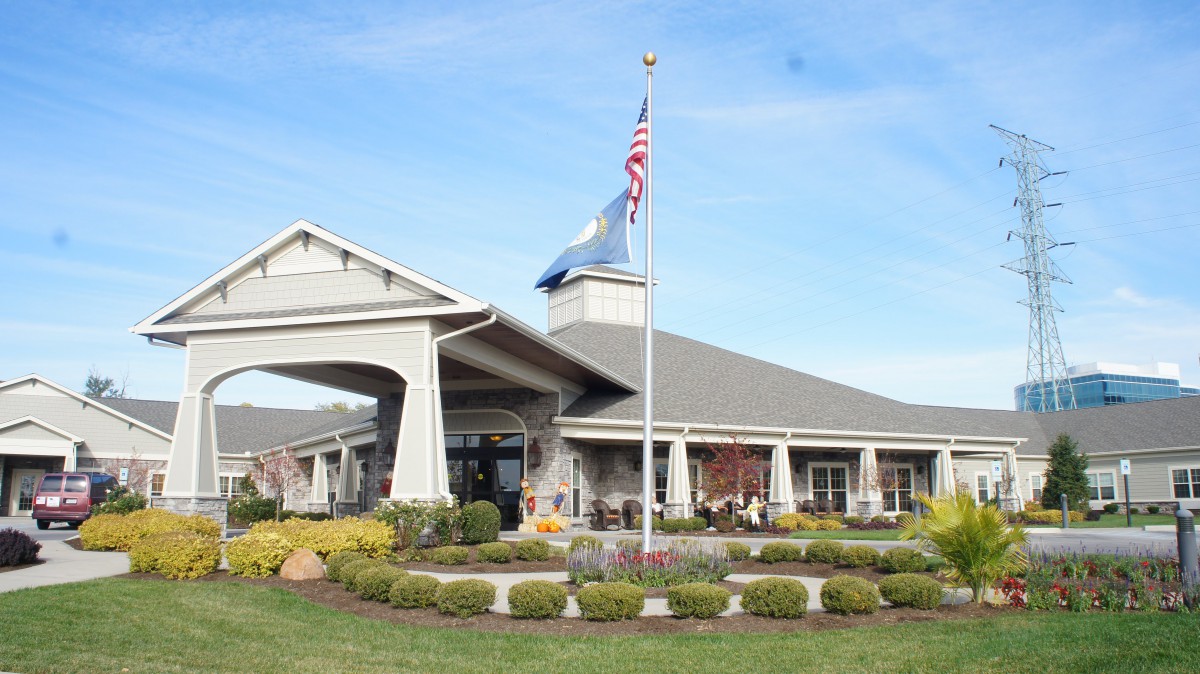 The image shows a large building with a sloped roof, featuring two flags flying in front and surrounded by neatly landscaped gardens.