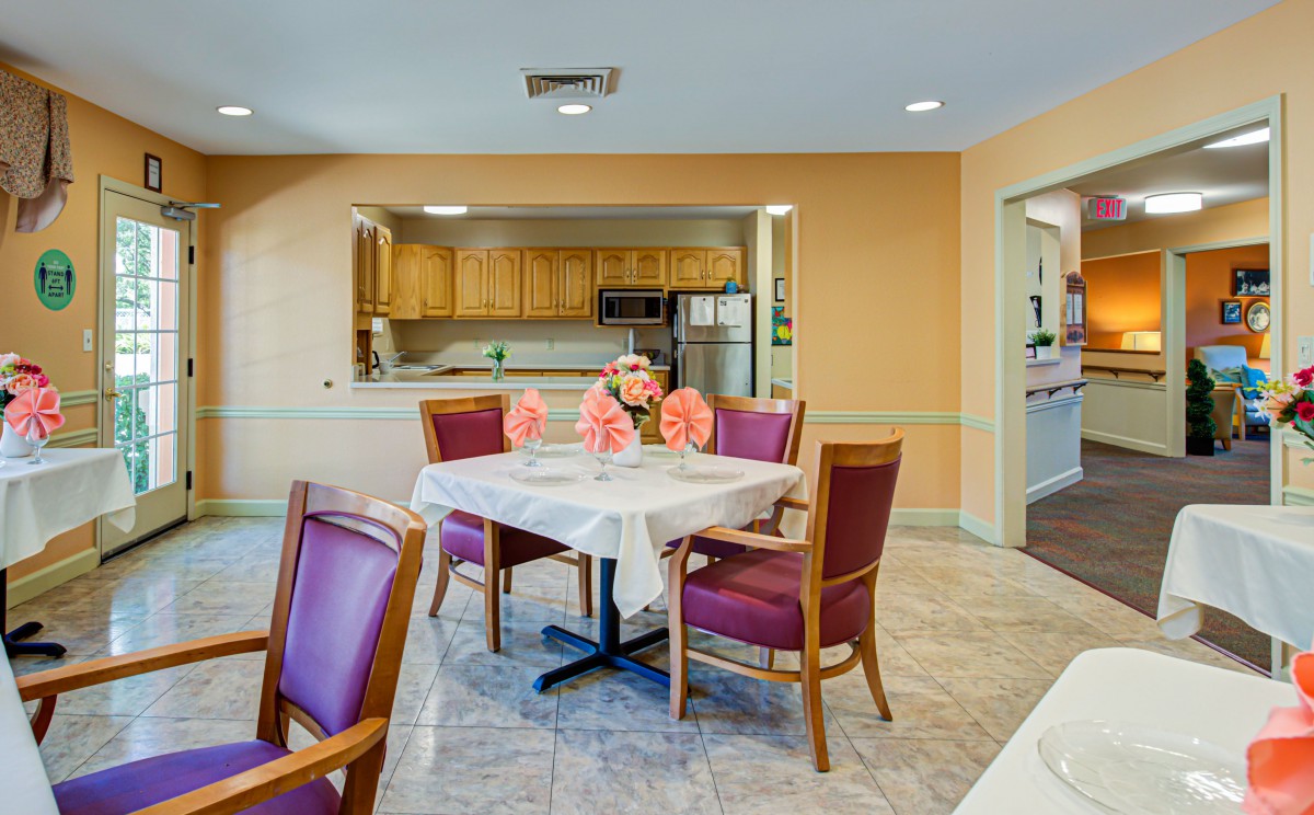 A cozy dining area with a table set for four, adorned with flowers, adjacent to a kitchen.
