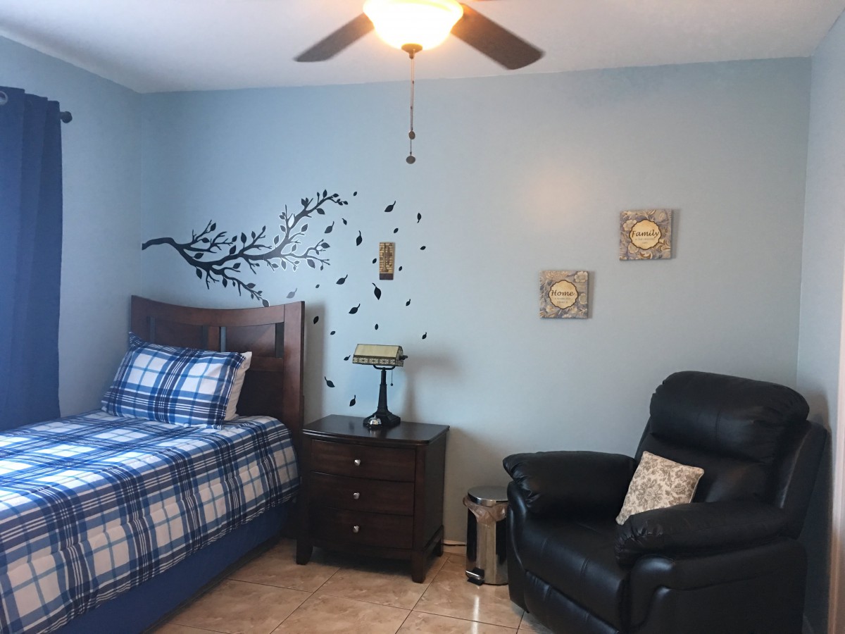 A cozy bedroom featuring a bed with checkered bedding, a nightstand with a lamp, and a comfortable armchair. The wall is decorated with artistic decals and framed pictures.