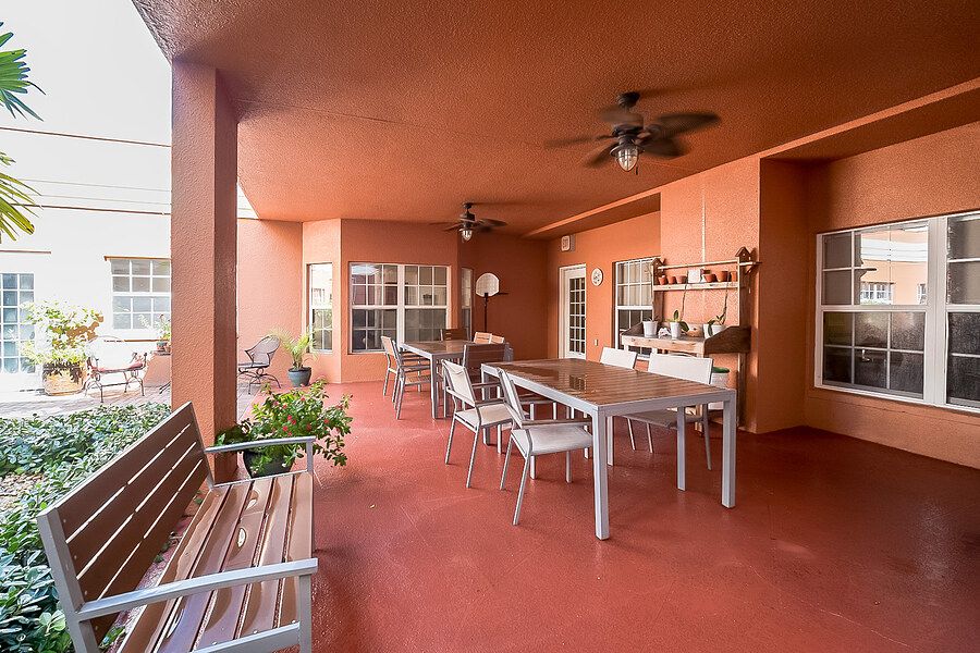 A spacious patio with outdoor seating and dining area under a terracotta-colored roof, featuring plants and fans for comfort.