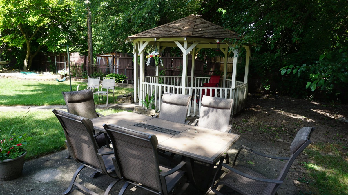 A tranquil garden scene featuring a gazebo surrounded by lush greenery and outdoor dining furniture on a patio.