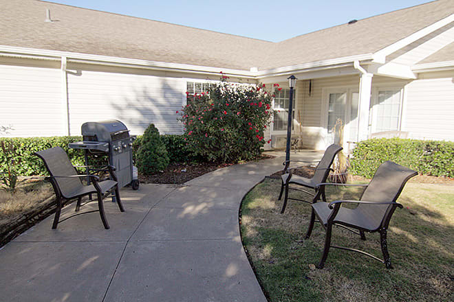 A cozy outdoor patio area with chairs, a grill, and a garden pathway in front of a house.