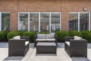 A modern outdoor patio setup with wicker chairs, sofas, and a coffee table in front of a brick wall with large windows and shrubbery.