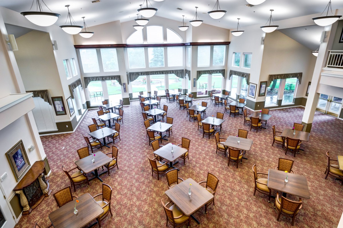 An empty dining hall with neatly arranged tables and chairs, featuring large windows and overhead lights.
