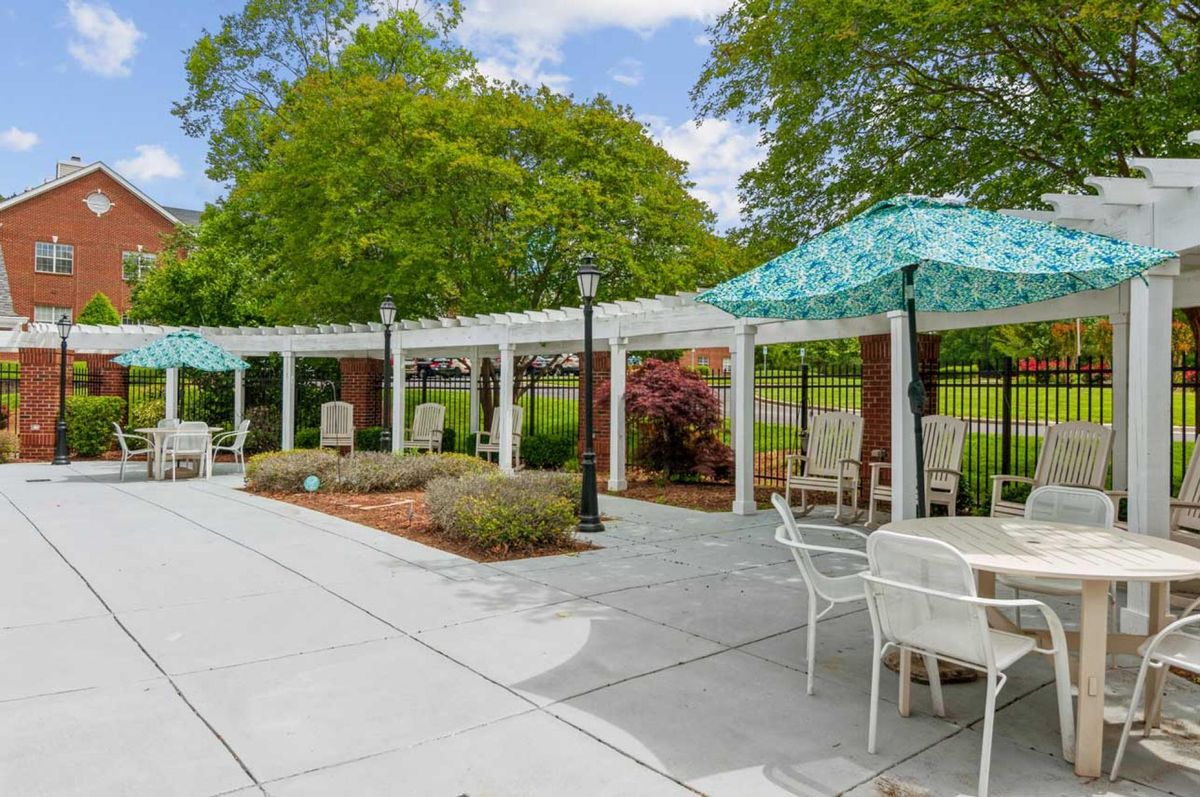 A serene patio area with a pergola, surrounded by lush greenery and furnished with tables and chairs.