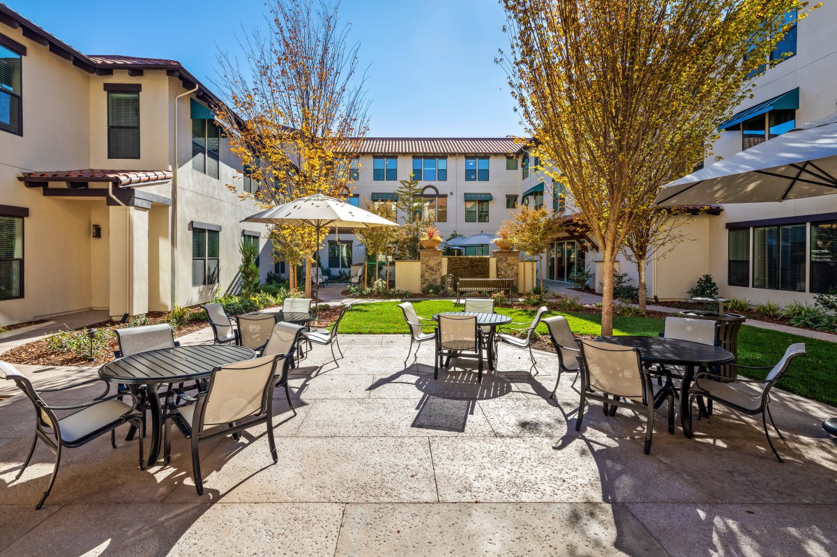 A sunny courtyard with outdoor seating and tables, surrounded by a building and trees.