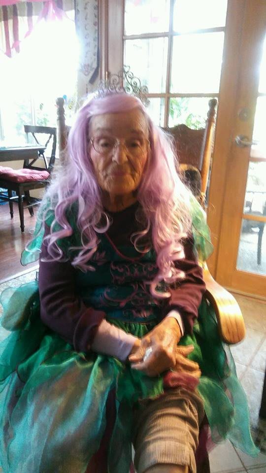 Elderly woman with purple wig sitting indoors.