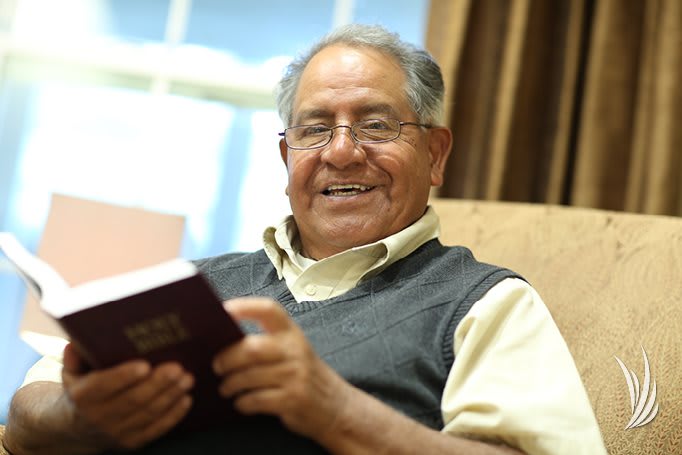 An elderly man smiling while reading a book indoors.