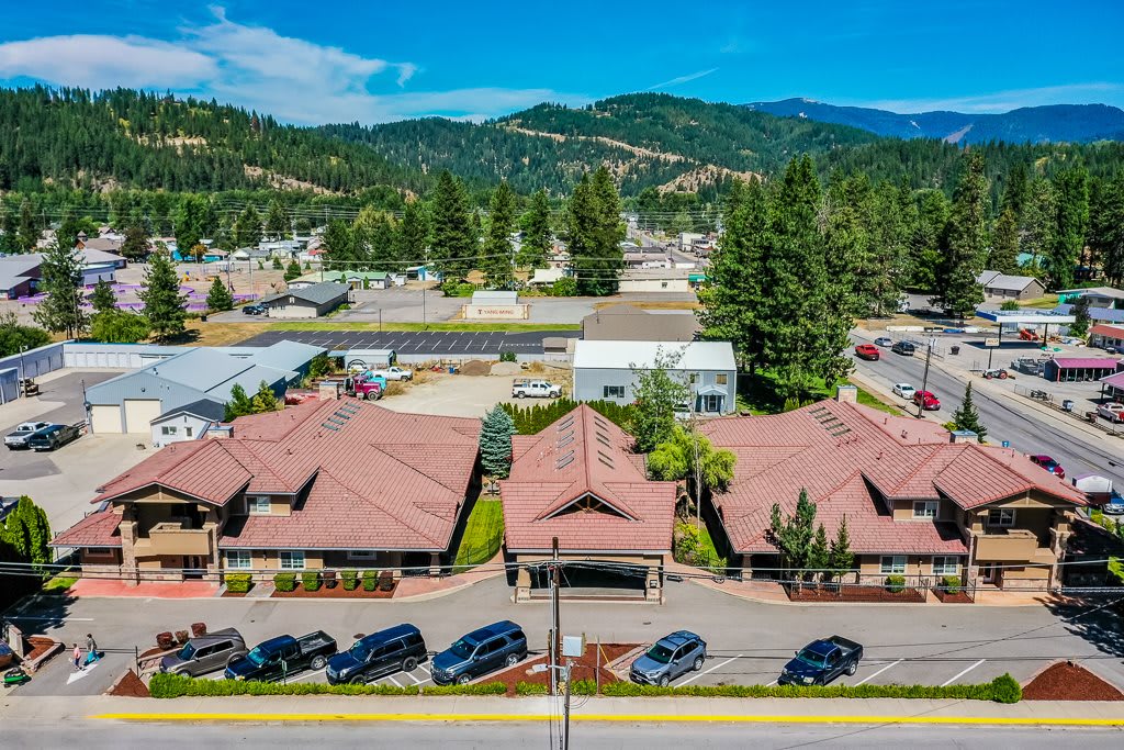 Aerial view of a complex with several buildings surrounded by mountains and trees, with cars parked along a road.