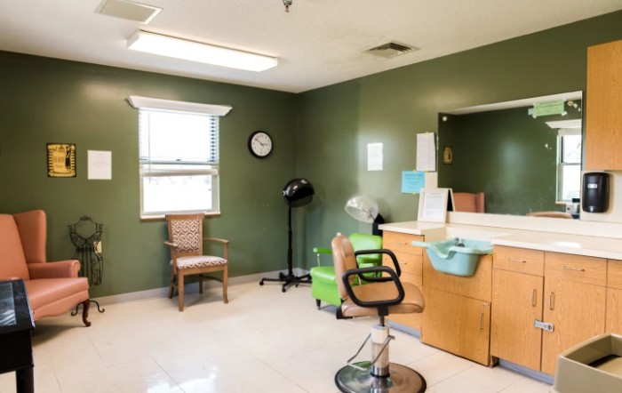 An empty hair salon room with green walls, featuring a styling chair, mirror, and salon equipment.