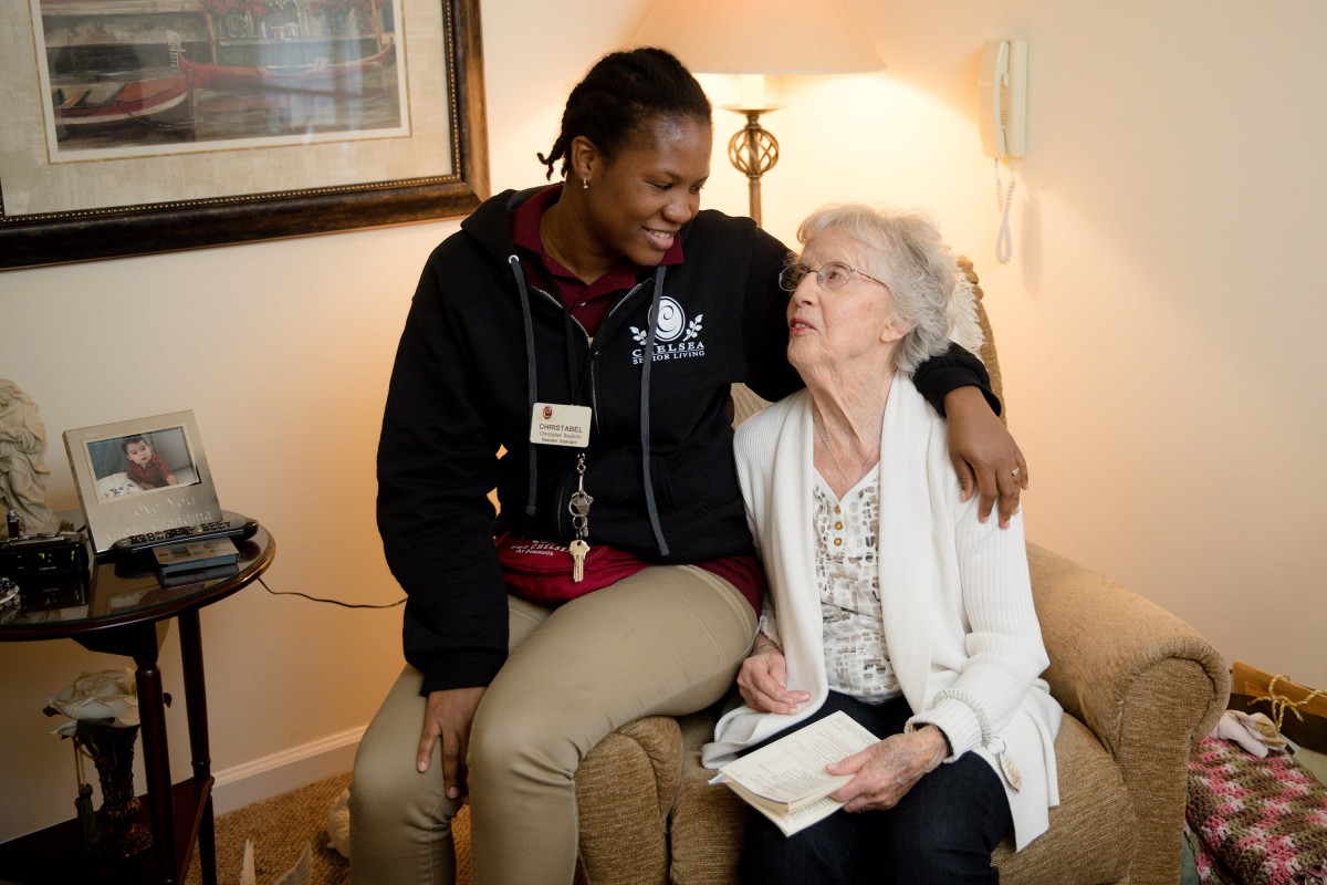 An elderly woman and a caregiver share a warm moment together, smiling in a comfortable setting.