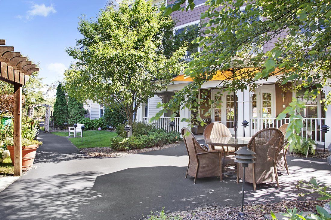 A charming outdoor patio with wicker furniture, surrounded by a lush garden and trees under a sunny sky.
