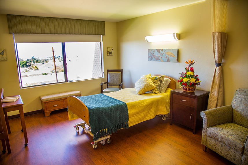A well-lit hospital room with a single bed, a chair, and a bouquet of flowers on a nightstand.