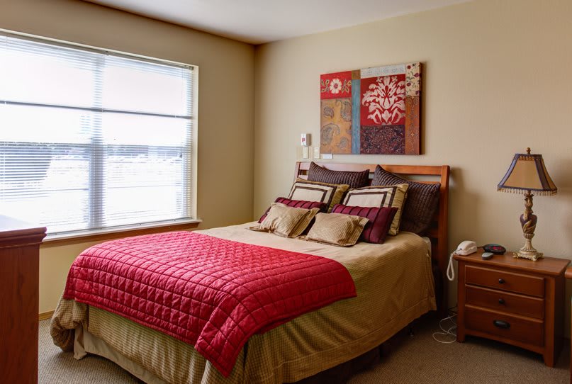 A cozy bedroom with a neatly made bed, featuring red and beige bedding and a decorative wall art piece.