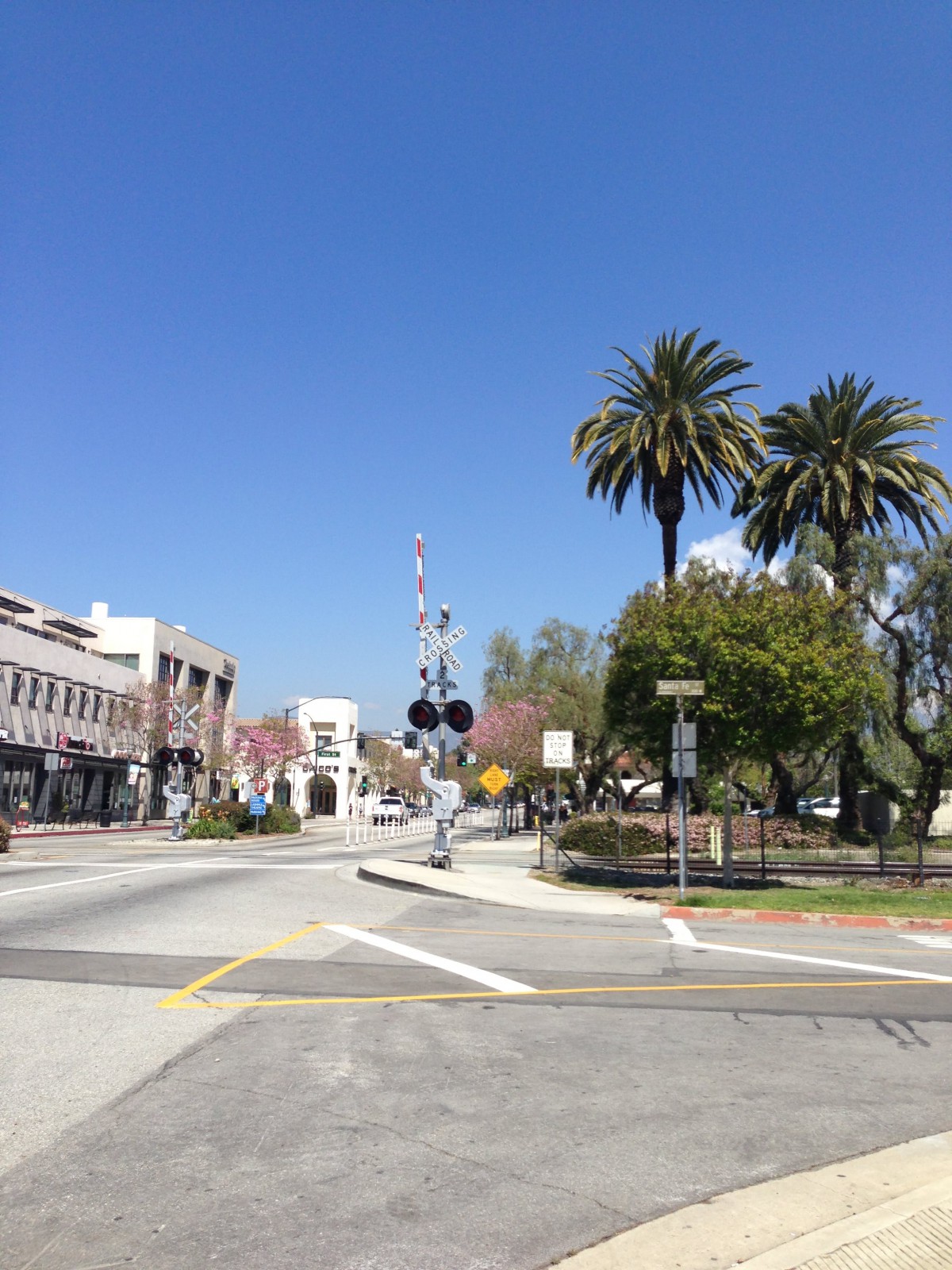 A sunny street scene with palm trees, buildings, and a railroad crossing.