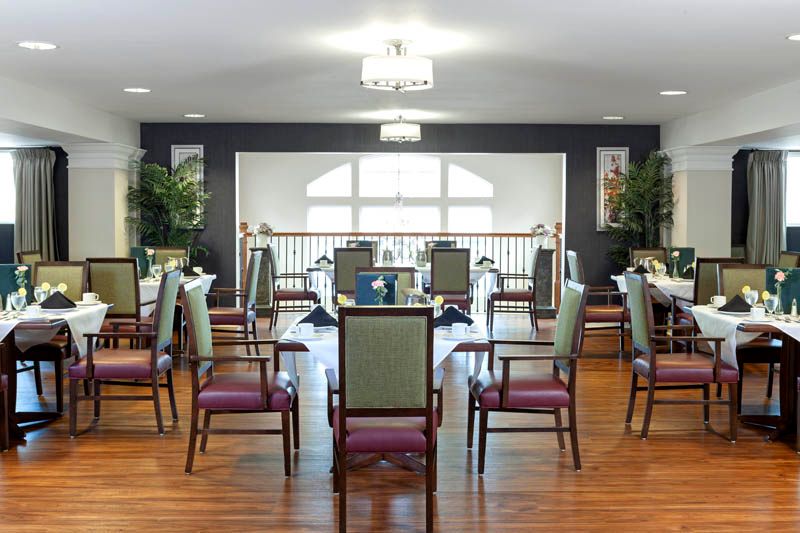 A spacious dining room with neatly arranged chairs and tables, set on a polished hardwood floor, accented by large windows and decorative plants.