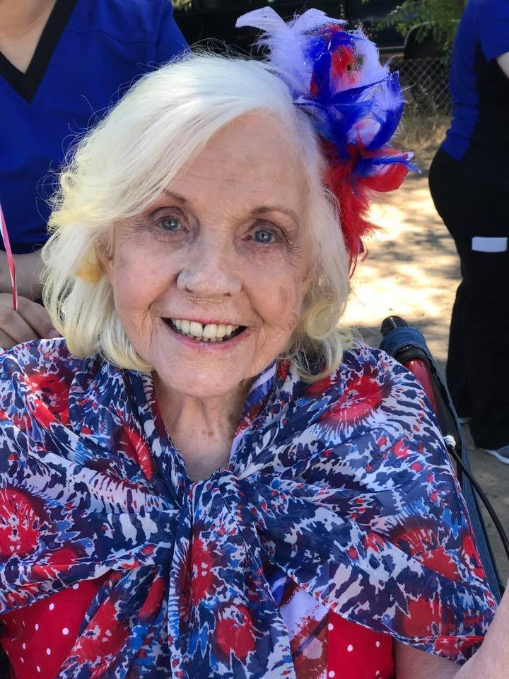 An elderly woman smiling outdoors, wearing a floral shawl in red, white, and blue.