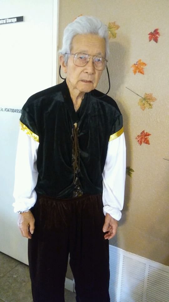 An elderly person dressed in a costume stands indoors with autumn-themed decorations on the wall.