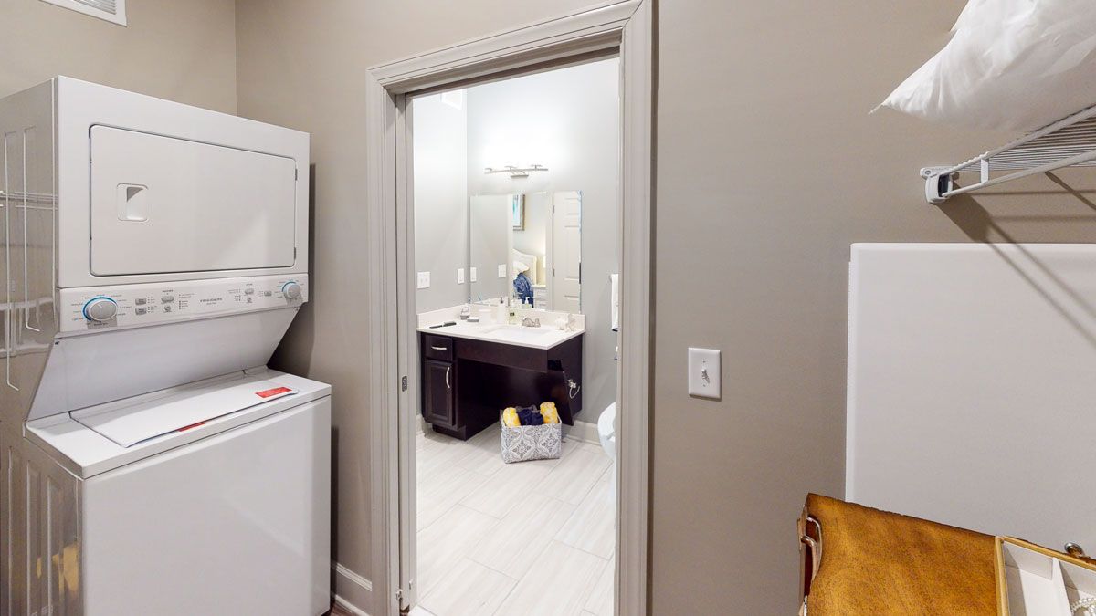 A view of a bathroom seen through an open door from a laundry area with a stacked washer and dryer.