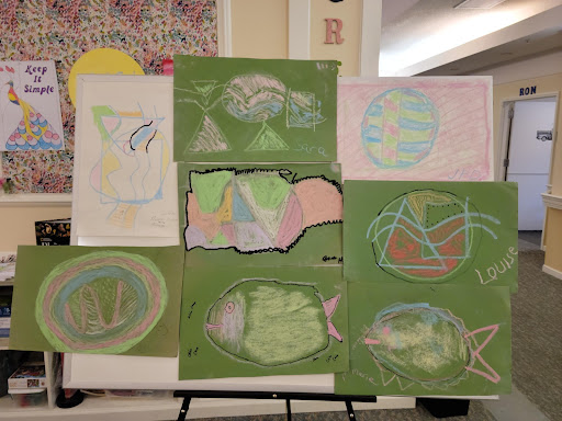 A collection of colorful children's artwork displayed on a board, featuring various abstract designs against green backgrounds.