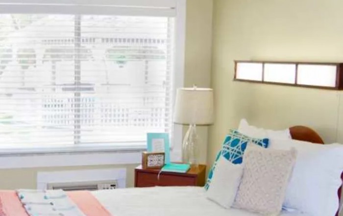 A cozy bedroom with a neatly made bed, decorative pillows, and a lamp on the nightstand beside a window.