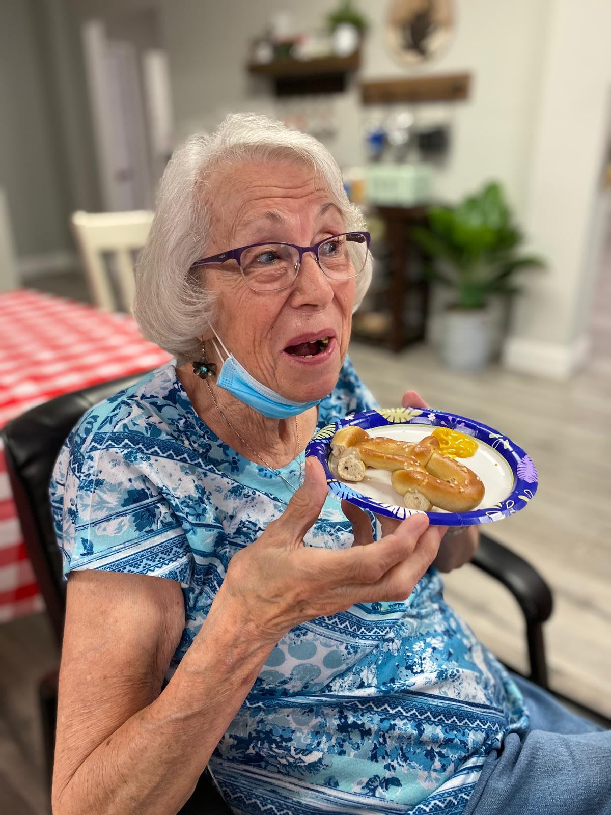 An elderly woman enjoys a meal indoors while wearing a mask under her chin.