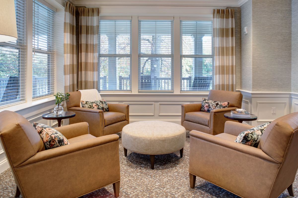 A cozy living room with comfortable chairs arranged around a central ottoman, large windows, and stylish curtains.