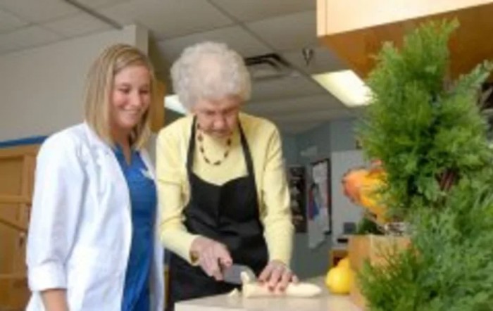 An elderly woman slices food alongside a young woman in a kitchen setting.