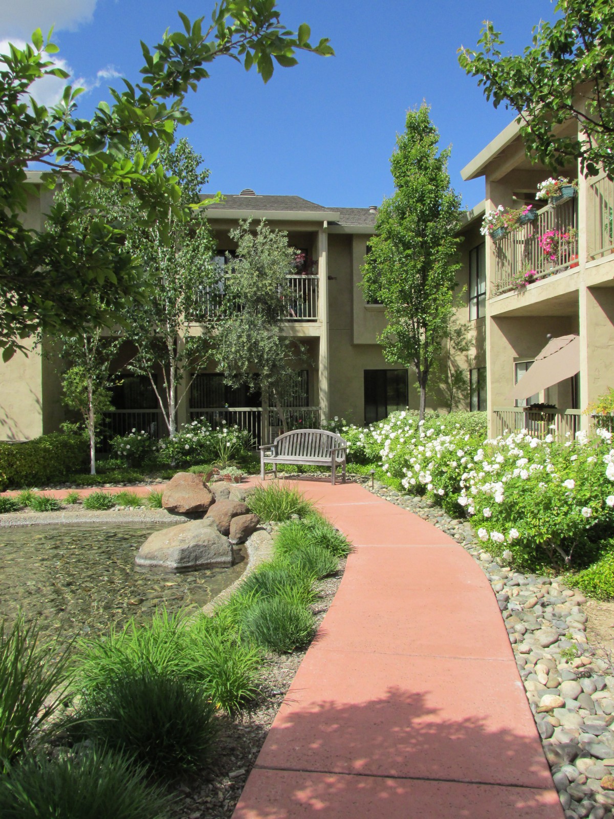 A serene apartment complex with a garden path leading to a pond, surrounded by lush greenery.