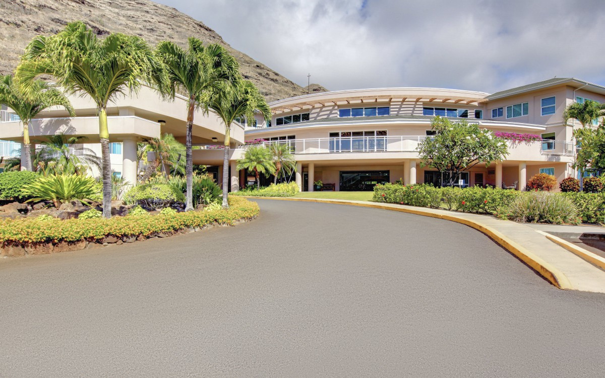 A modern building surrounded by palm trees and landscaped gardens under a partly cloudy sky.