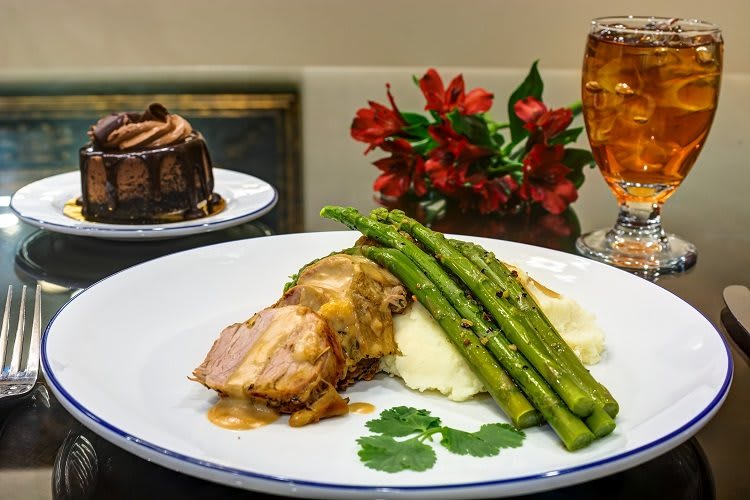 A delicious dinner plate featuring pork with gravy, asparagus, and mashed potatoes, accompanied by a dessert cake and a drink.