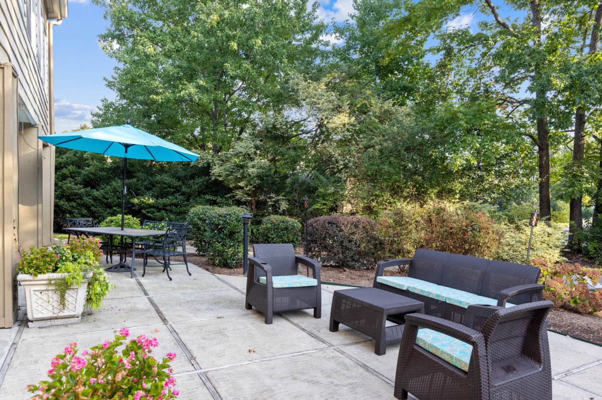 A cozy patio with outdoor seating, surrounded by lush greenery and shaded by an umbrella.