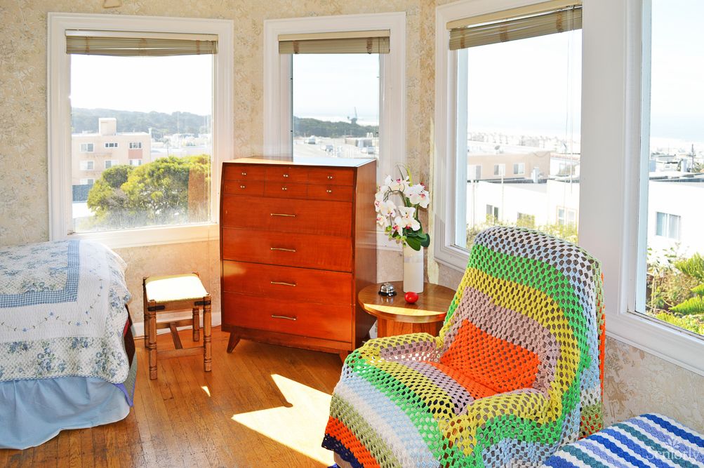 A cozy living room with an armchair covered in a colorful blanket, wooden furniture, and large windows offering a scenic view.