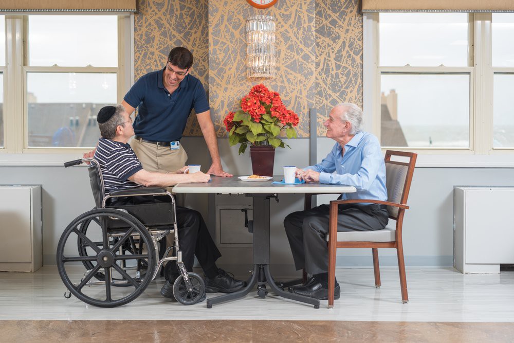 A caregiver interacting with two elderly individuals at a table in an indoor setting.